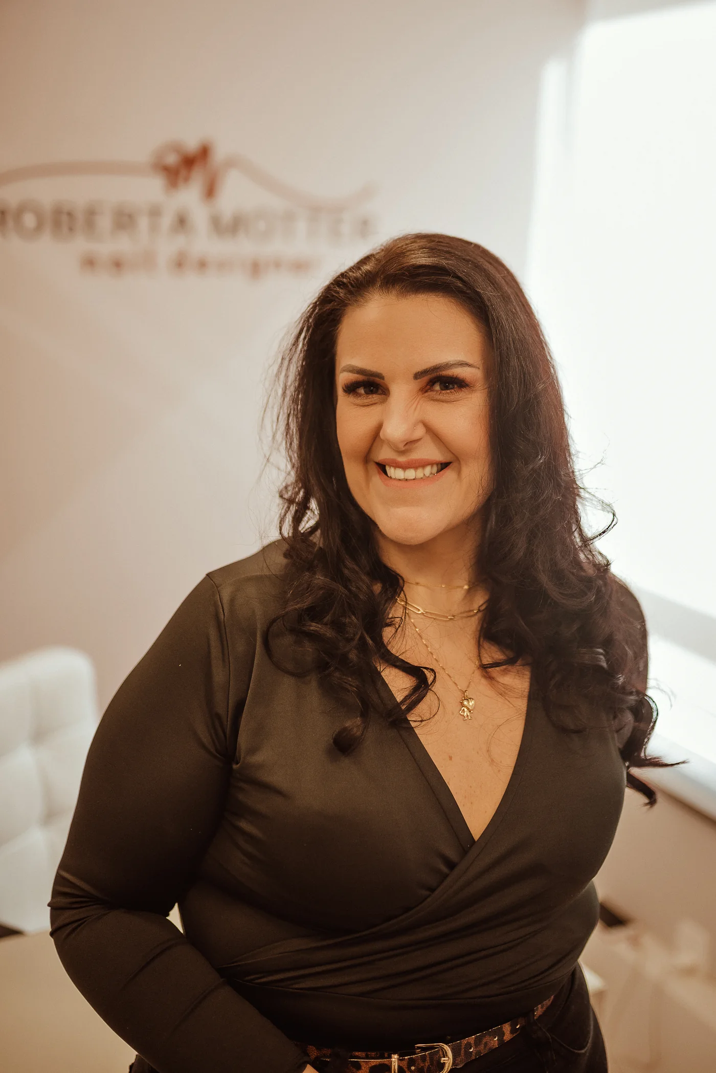 Roberta Motter, nail designer, sorrindo em frente à parede com o logotipo do estúdio